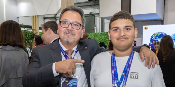 Two men posing for a photo at a conference; one in a suit and glasses points at the younger man who smiles, with lanyards visible in a busy expo background.