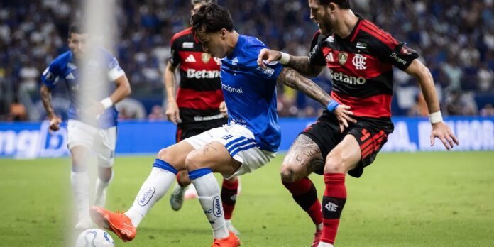 Soccer players in blue and red-black uniforms compete for the ball during a match, near the sideline on a green pitch.
