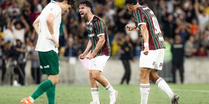 Three soccer players in maroon–green striped jerseys celebrate a goal on the field as the crowd cheers in the stands.