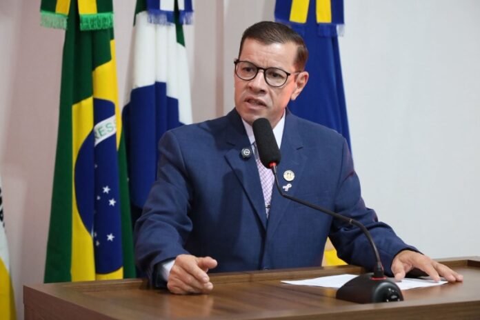 Man in a blue suit and glasses speaks into a microphone at a wooden podium, flags behind him.
