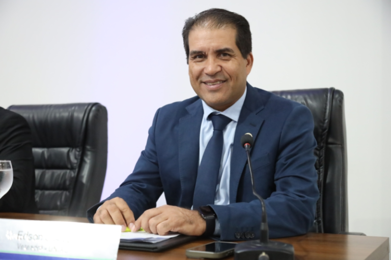 Man in a navy suit and tie sits at a conference table, smiling, with a microphone in front of him.