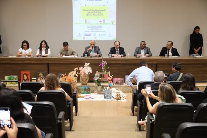 Panel of eight people seated at a long table on a stage, presenting to an audience with a poster projected behind them.
