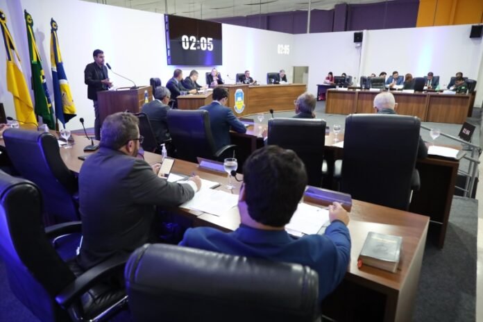 Formal council meeting with attendees around a U-shaped conference table; speaker at podium. Flags line the wall on the left.