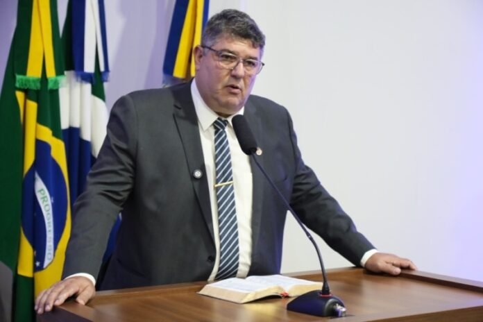 Man in a dark suit and striped tie speaking at a podium with a microphone, open book in front, Brazilian flags in the background