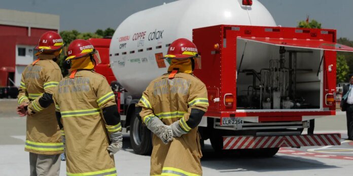 Three firefighters in tan turnout gear and red helmets stand with their backs to the camera, facing a red fire truck with an open equipment compartment and a large white tanker behind them on a tarmac.