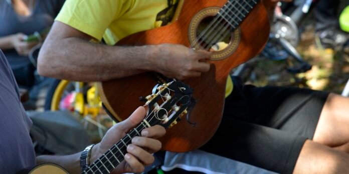 Two people playing acoustic guitars outdoors, close-up on hands and fretboards during a casual jam session in daylight, with one player wearing a yellow shirt.