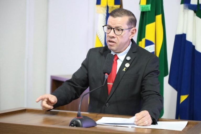 Man in a dark suit and red tie speaks at a podium with a microphone, gesturing with his hand in front of flags in the background.