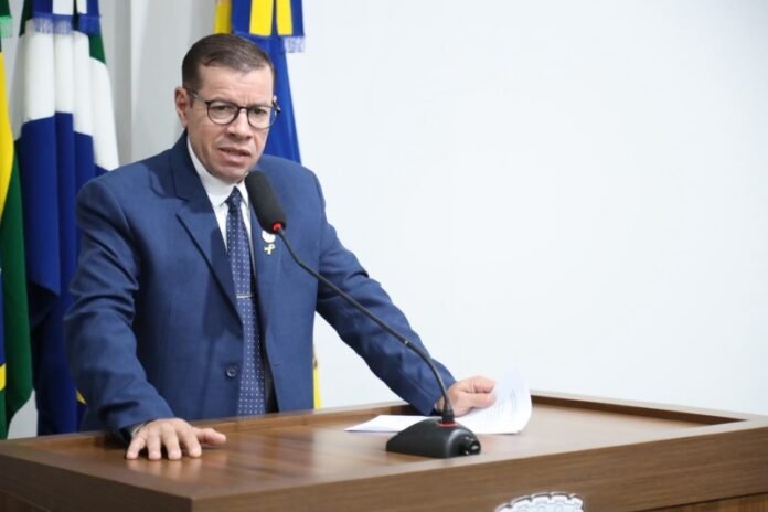 Man in a navy suit and glasses speaks at a wooden podium with a microphone, flags in the background.