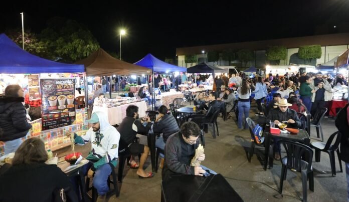 Night market with colorful tents, food stalls, and people sitting at plastic tables eating in the evening light.
