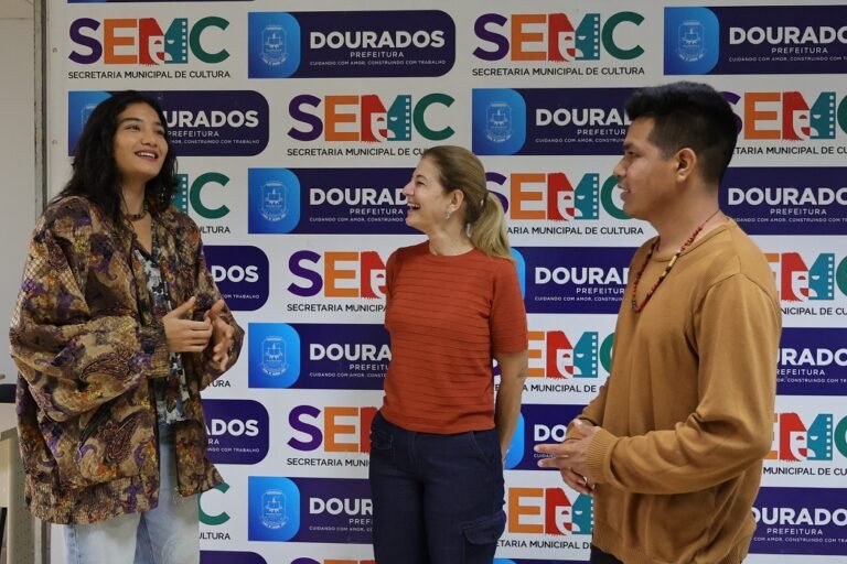 Three people stand and smile while conversing in front of a backdrop covered with SEMAC and Dourados Prefeitura logos.
