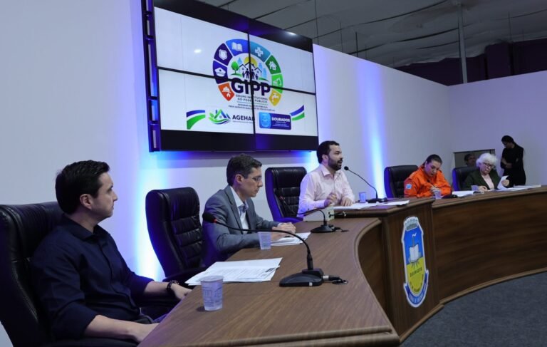 Panel of seven people seated at a curved wooden dais in a conference room, microphones in front, a large screen with a colorful logo behind them and blue lighting along the walls.