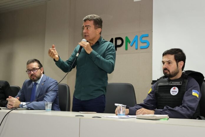 Man speaks into a handheld microphone at a panel, gesturing with his left hand; two men sit beside him in a conference room with a logo on the wall behind them.