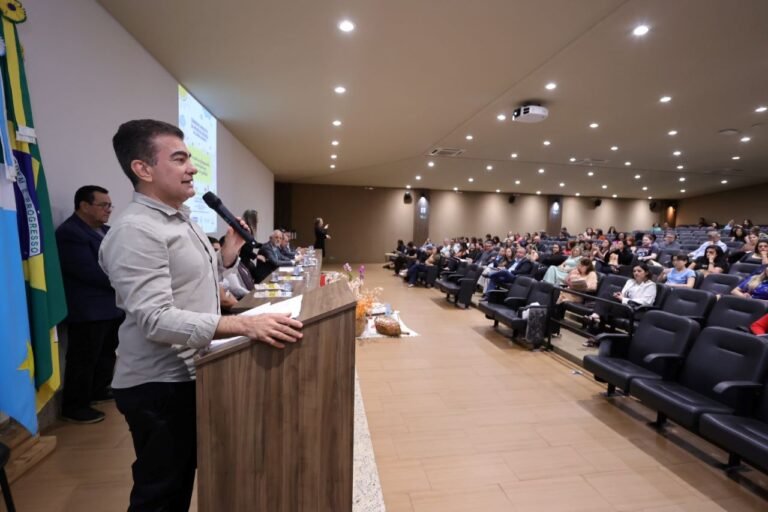Man at a wooden podium speaking into a microphone to a seated audience in a conference hall, with flags on the left.