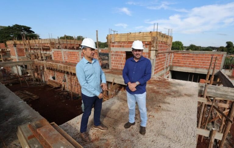 Two construction workers wearing white hard hats stand on a concrete ledge above a brick building under construction, with scaffolding around them.