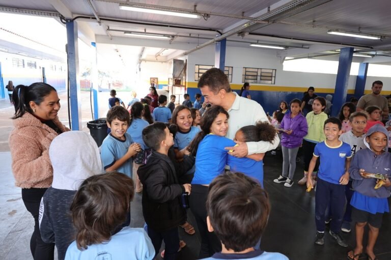 A man in a light shirt hugs a girl in blue as smiling students surround them in a covered school courtyard.