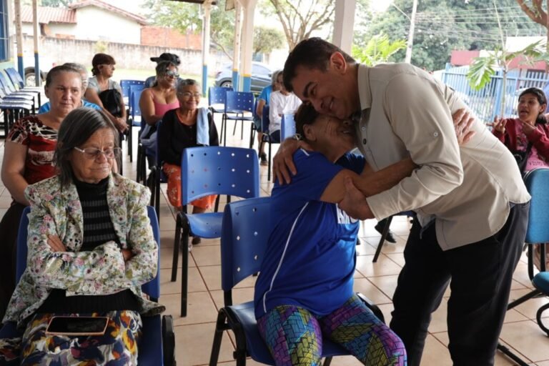Man helps an elderly woman dance and hug at a community gathering, with other seniors seated nearby.