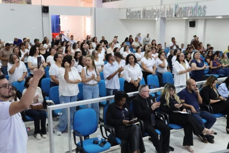Large crowd in a municipal chamber, with many people standing and clapping, seated on blue chairs; signage reads 'Câmara Municipal de' in the background.