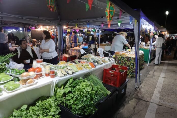 Night market scene with colorful tents, string lights, and tables overflowing with fresh vegetables and prepared foods at night.