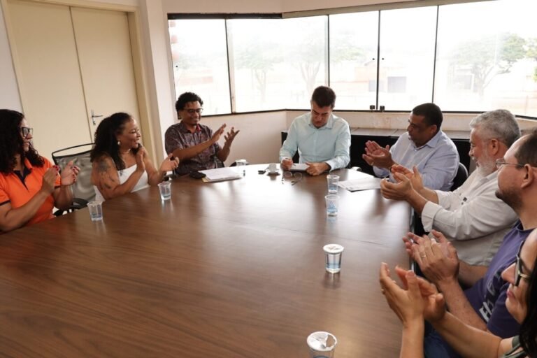 Group of coworkers around a long conference table, clapping in a bright office meeting room.