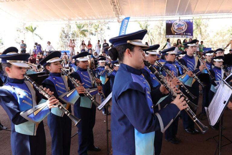Young band in blue uniforms playing clarinets at an outdoor event under a shade canopy, with spectators in the background.