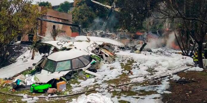 Wrecked small airplane fuselage in a foam-covered crash site, debris scattered with firefighters in the background.