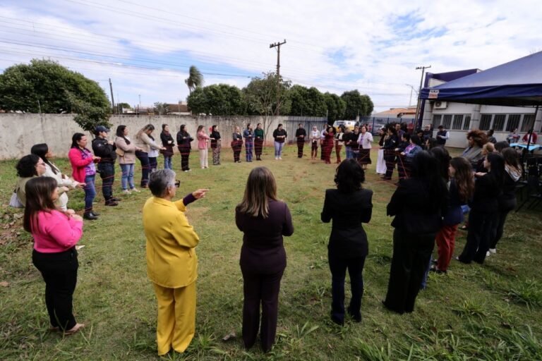 People stand in a large circle on a grassy lot during an outdoor group activity, facing inward toward the center.