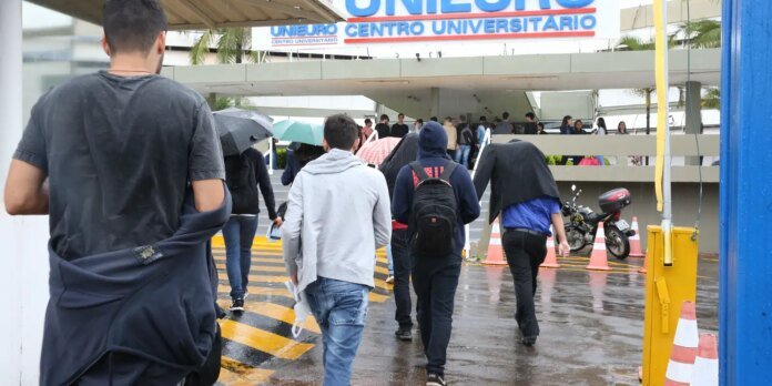 Group of students with backpacks walking toward a university entrance on a rainy day, holding umbrellas under a covered walkway near a 'Centro Universitario' sign.