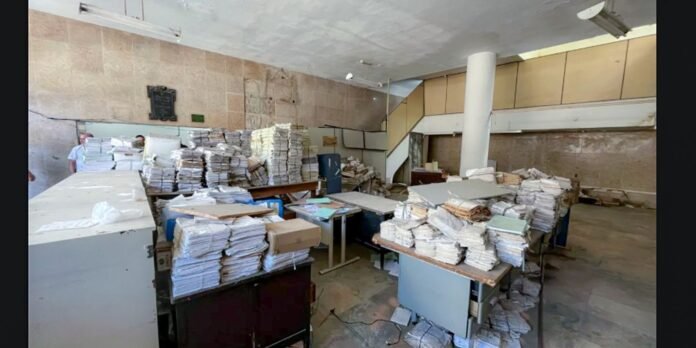 Cluttered office or archive room with numerous stacks of papers and folders piled on desks and shelves, creating a chaotic workspace.