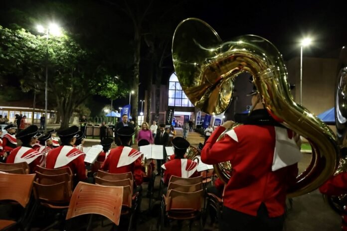 Nighttime outdoor brass band performance; musicians in red uniforms seated with large shiny tubas in the foreground and a lit building in the background.
