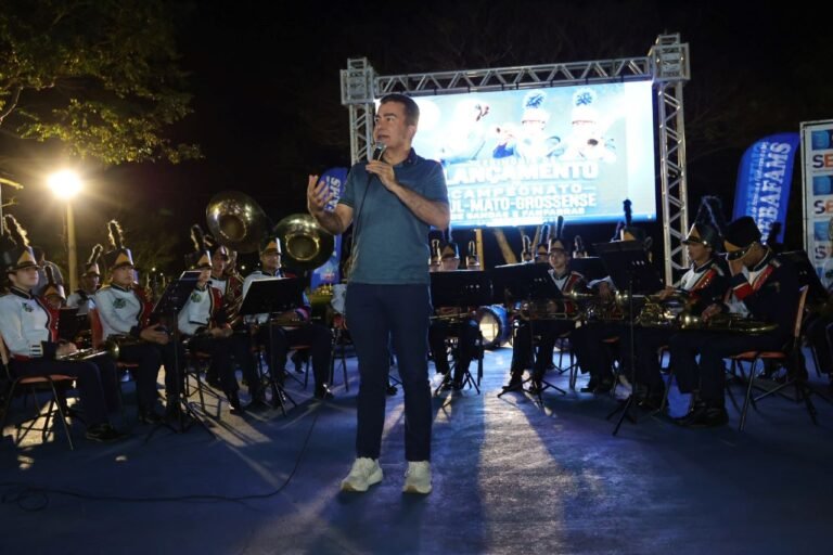 Man speaking into a microphone in front of a seated brass band on an outdoor stage at night.