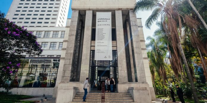 Front view of a stone library entrance with a tall vertical banner and people on the steps, surrounded by palm trees and flowering bushes.