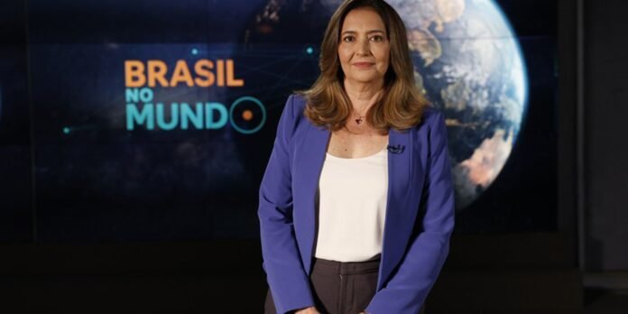 News anchor standing in a studio with a large Earth backdrop and 'BRASIL NO MUNDO' on screen, wearing a purple blazer and white top.