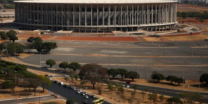A round stadium with numerous vertical concrete columns, viewed from above, surrounded by roads, parking lots, and sparse trees on a dry day.