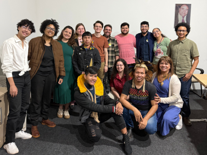 Diverse group of about 15 adults posing together in a room, some standing and two seated on the floor, smiling at the camera.
