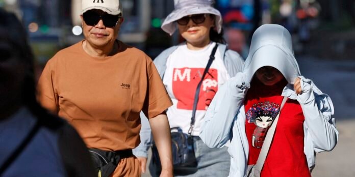 Three people walk together on a sunny street: a man in an orange t‑shirt with a cap and sunglasses, a woman in a white hat, and a person in a light jacket adjusting the hood.