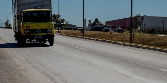 Front view of a yellow delivery truck driving toward the camera on a wide road near an industrial area with parked vehicles in the distance.