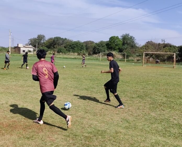A group of young players on a grassy soccer field, one in a pink jersey chasing a ball while others watch or run toward play in the background.