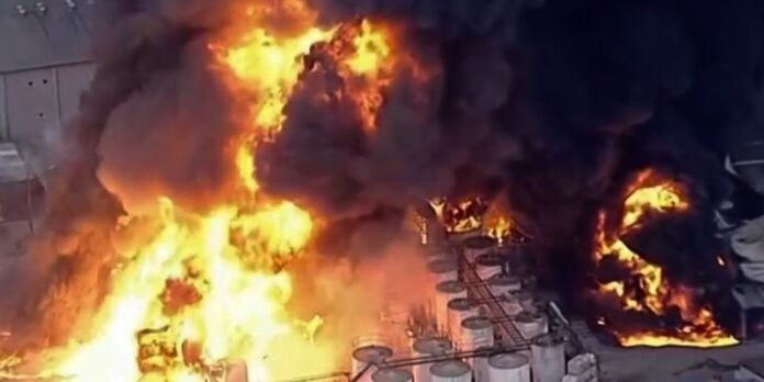 Industrial fire with large orange flames and thick black smoke at a facility, several cylindrical tanks nearby on fire and burning debris nearby.