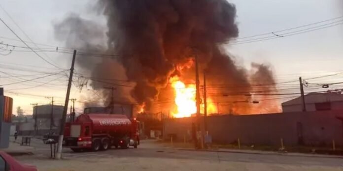 Building on fire with large flames and thick black smoke; a fire truck is on the street nearby while utility poles loom overhead.