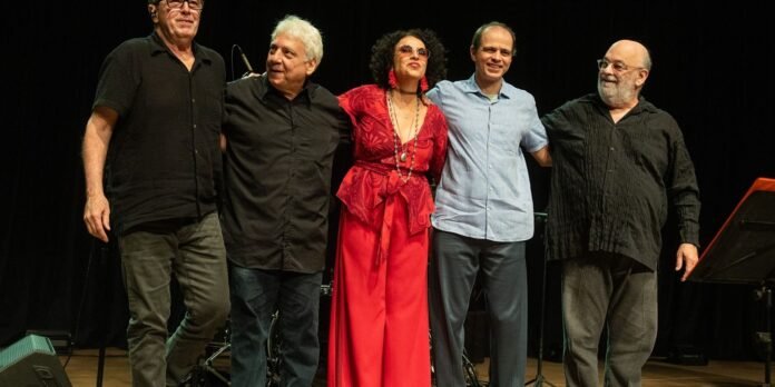 Five adults stand on a stage with arms around each other, smiling after a performance, wearing casual to semi-formal clothing including a red dress and black shirts.