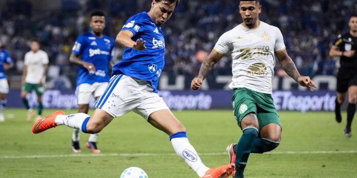 Two soccer players compete for the ball during a match: a blue-jersey player kicking while a white-green-jersey defender approaches, ball on the ground nearby.