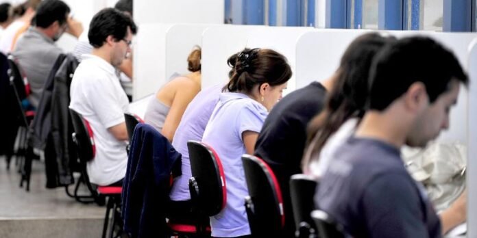 People seated at individual desks in a row of cubicles inside a bright office, working at computers/with papers.