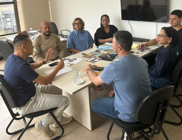 Group of eight adults seated around a conference table in a bright office, discussing with brochures and papers spread out. A wall-mounted screen is in the background.