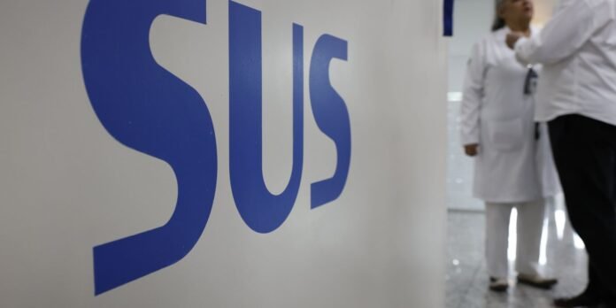 Close-up of large blue letters painted on a white wall with medical staff in white coats walking in a hospital corridor in the background.
