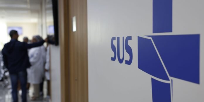 Close-up of a blue SUS logo on a wall, with a blurred hospital corridor and people in the background.