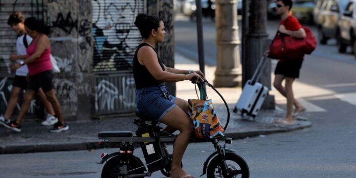 Woman rides a compact black bike on a city street with a colorful tote bag in front. Graffiti wall and pedestrians in background.