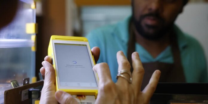Cashier uses a yellow handheld payment terminal at checkout, with a bearded worker in the background.