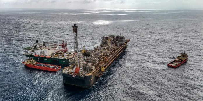 Large offshore drilling barge with complex rig equipment, flanked by two red tugboats in open sea against a cloudy sky