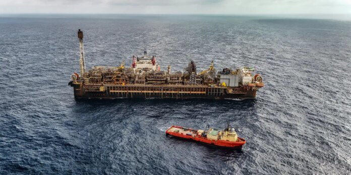 Offshore oil drilling platform on the ocean with a red support vessel nearby under a cloudy sky.