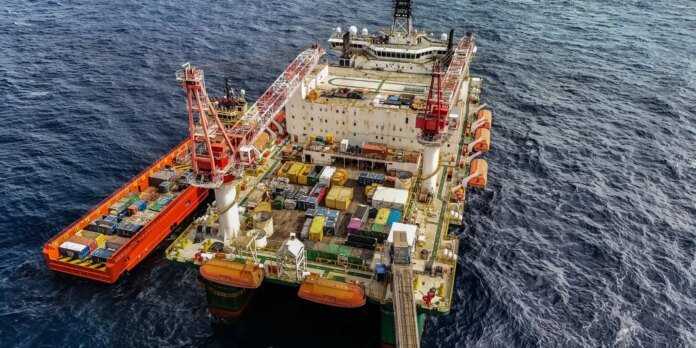 Aerial view of a large offshore support vessel with orange cranes and colorful cargo containers on the deck, sailing in dark blue sea.